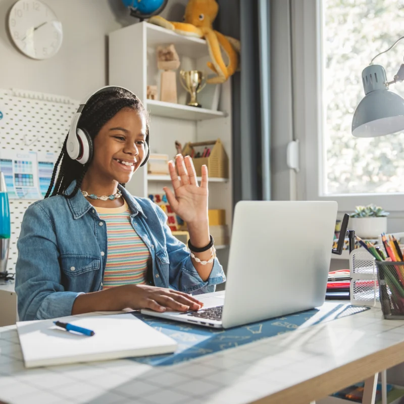 A smiling girl wearing headphones sits at her desk, waving at a laptop during a video call. Surrounded by notebooks, pencils, and a lamp in her bright room, she enjoys Online Learning Solutions for K-12.